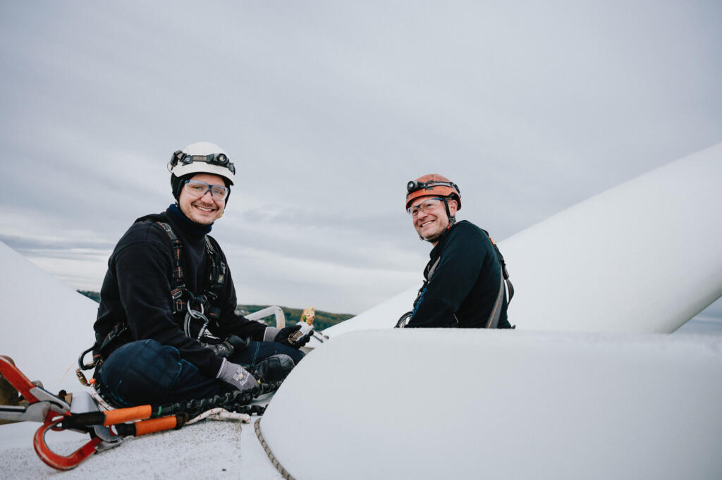 Zwei Personen in Kletterausrüstung sitzen auf einer Windkraftanlage und blicken lächelnd in die Kamera. Im Hintergrund sind Wolken zu sehen, die einen klaren Himmel über der Landschaft andeuten. Der Moment vermittelt Vertrauen und Teamarbeit im Bereich Employer Branding von Enertrag.