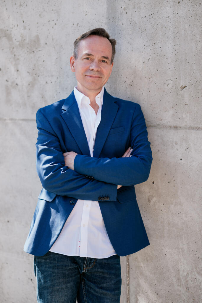 Ein Mann in einem blauen Blazer und weißem Hemd steht mit verschränkten Armen vor einer betonfarbenen Wand. Der Hintergrund ist neutral und betont die formelle Kleidung des Mannes, passend für Krebs & Krappen Business Portraits.