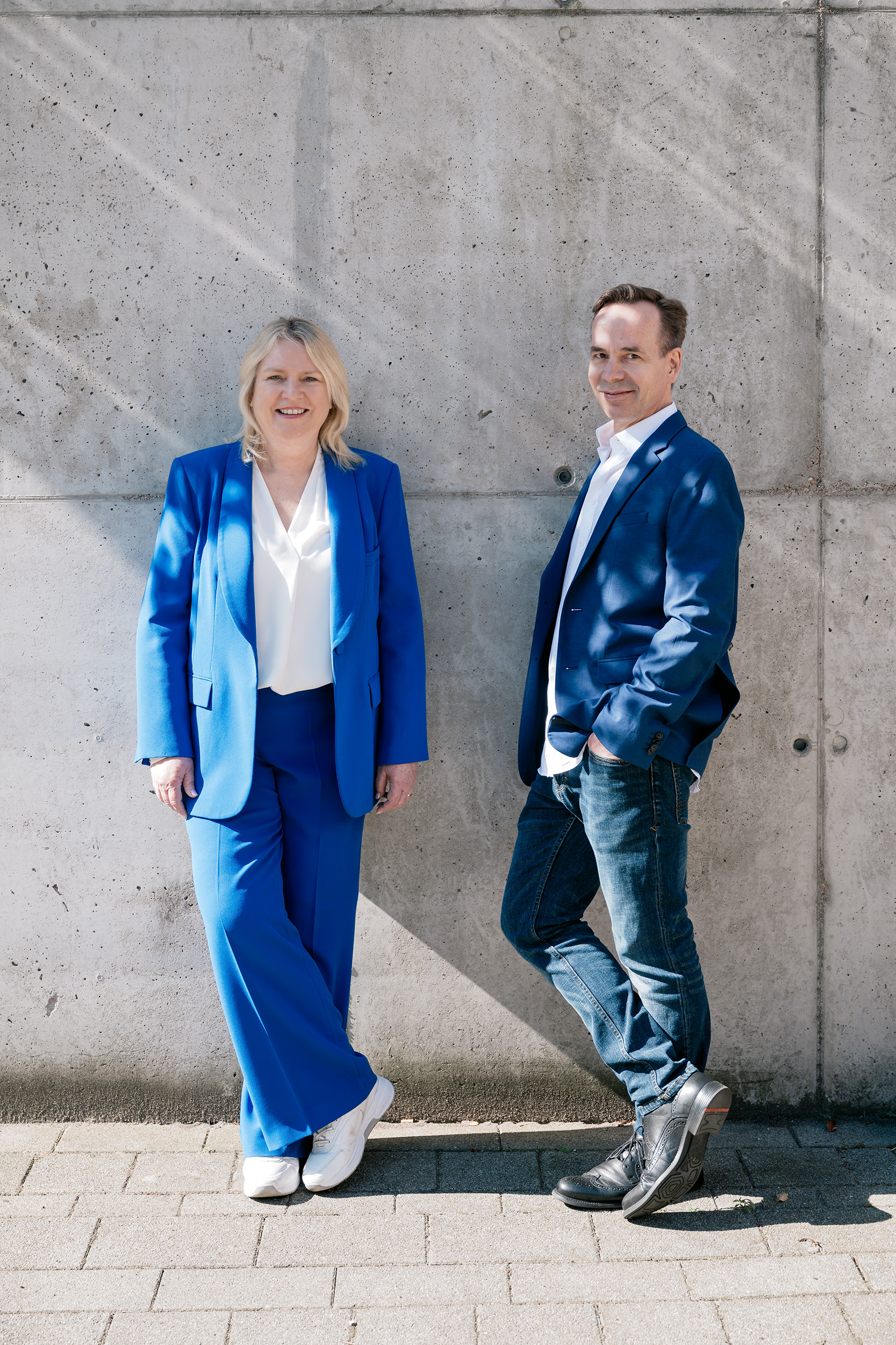 Zwei Personen in blauen Anzügen stehen lässig an einer Betonwand. Die Frau trägt eine blaue Bluse und Hose, der Mann ein blaues Sakko mit Jeans. Hintergrund zeigt Licht- und Schattenspiele.