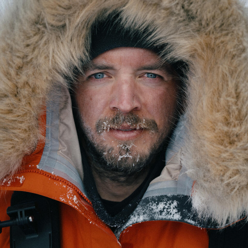 Ein Mann mit blauen Augen trägt eine orangefarbene Winterjacke mit einem Fellkragen. Sein Gesicht ist von Kälte und Schnee geprägt, während er in einer schneebedeckten Umgebung steht.