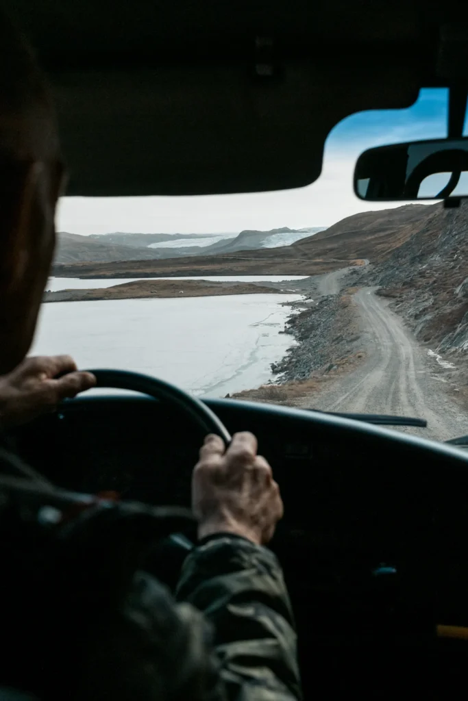 Ein Fahrer hält das Lenkrad eines Fahrzeugs, während er eine schmale, abgeschiedene Straße entlang einer Küste in einer bergigen Landschaft fährt. Die Umgebung zeigt Wasser und hügeliges Gelände, das für "The Greenland Project" relevant ist.