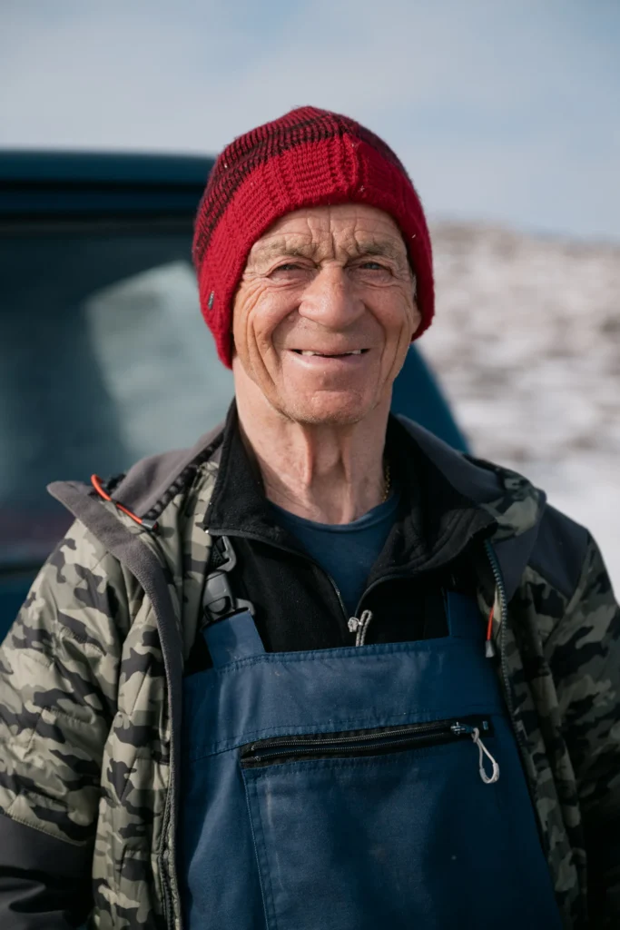 Ein älterer Mann mit einem roten Strickmütze und einem blauen Overall steht im Freien vor einem verschwommenen Hintergrund. Er lächelt und zeigt freundliche Züge.