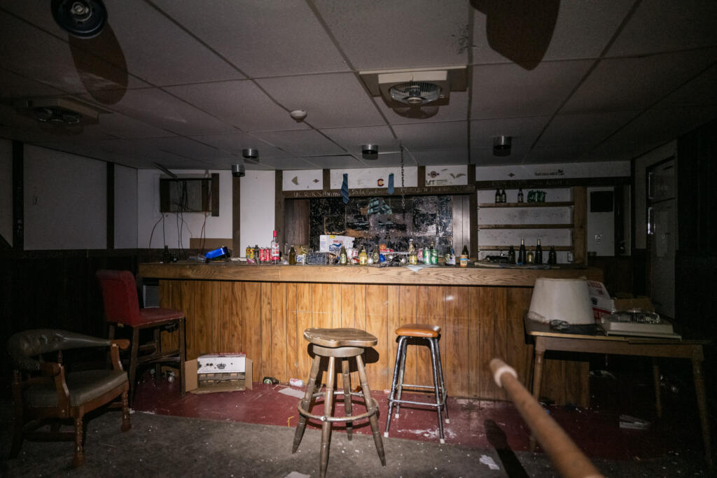 Ein verlassener Raum mit einer Holzbar, auf der sich leere Flaschen und Dosen befinden. Stühle sind unordentlich um die Bar angeordnet und der Boden zeigt Spuren von Verschmutzung. Das Licht ist schwach und die Wände sind dunkel.