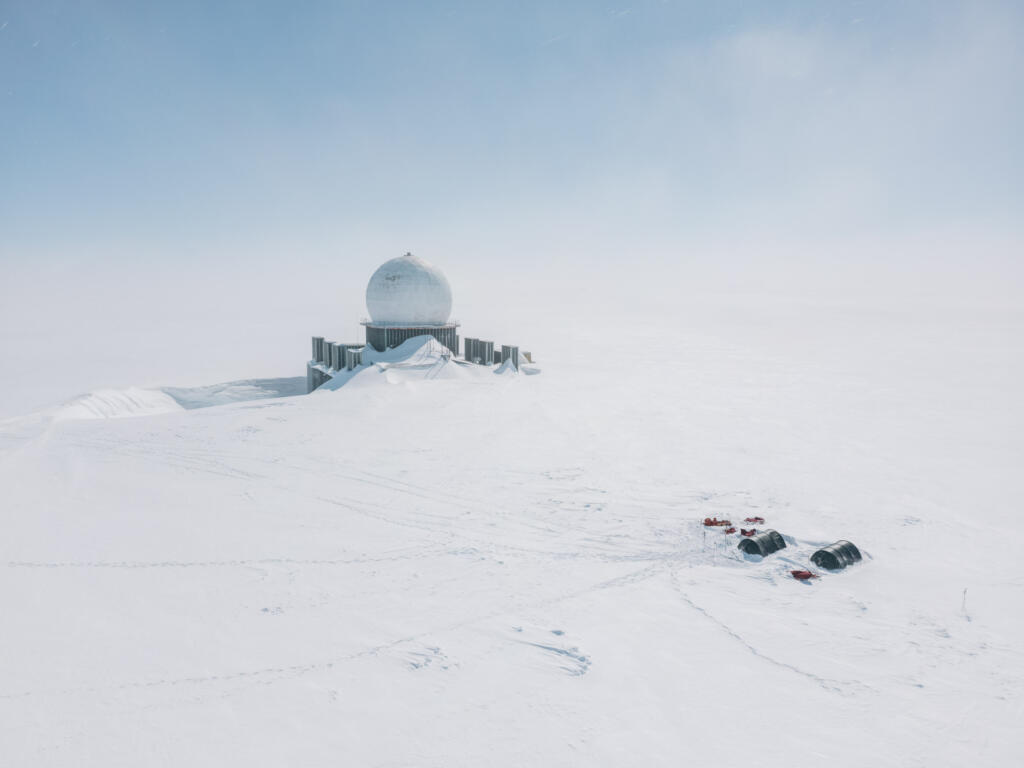 Eine von Schnee bedeckte Forschungsstation mit einer kuppelförmigen Struktur, umgeben von einer schneebedeckten Landschaft unter einem klaren Himmel. Im Vordergrund sind Zelte und Fahrzeuge sichtbar.