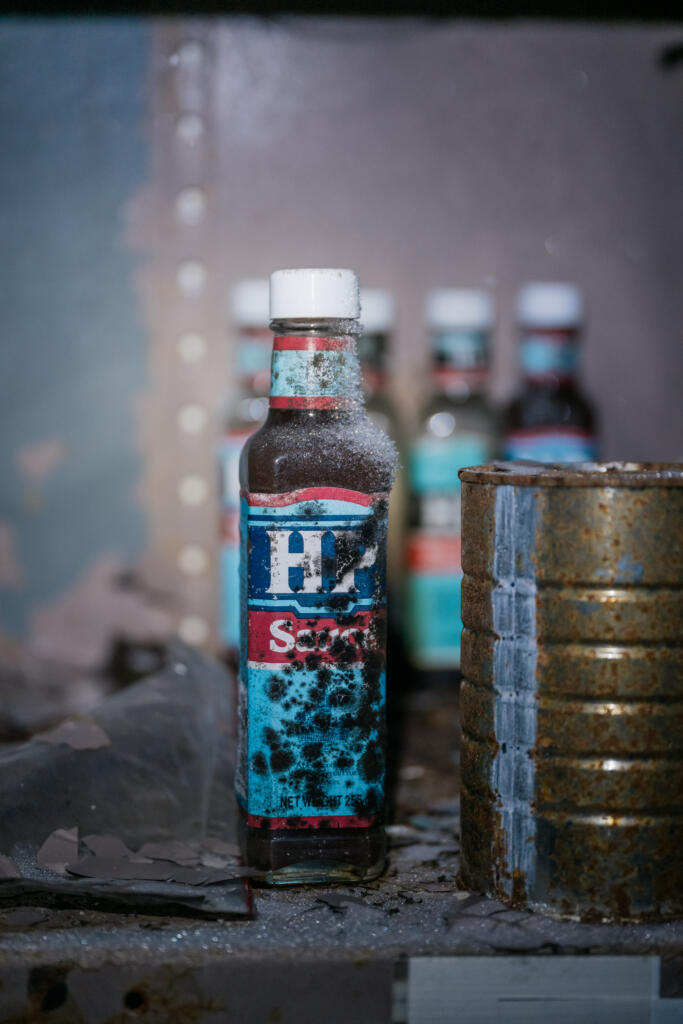 Eine verschmutzte Flasche mit blauer Flüssigkeit, beschriftet mit "H2O", steht auf einem Regal. Im Hintergrund sind weitere Flaschen und eine rostige Dose sichtbar. Die Umgebung wirkt vernachlässigt und staubig. #Dye-2-greenland
