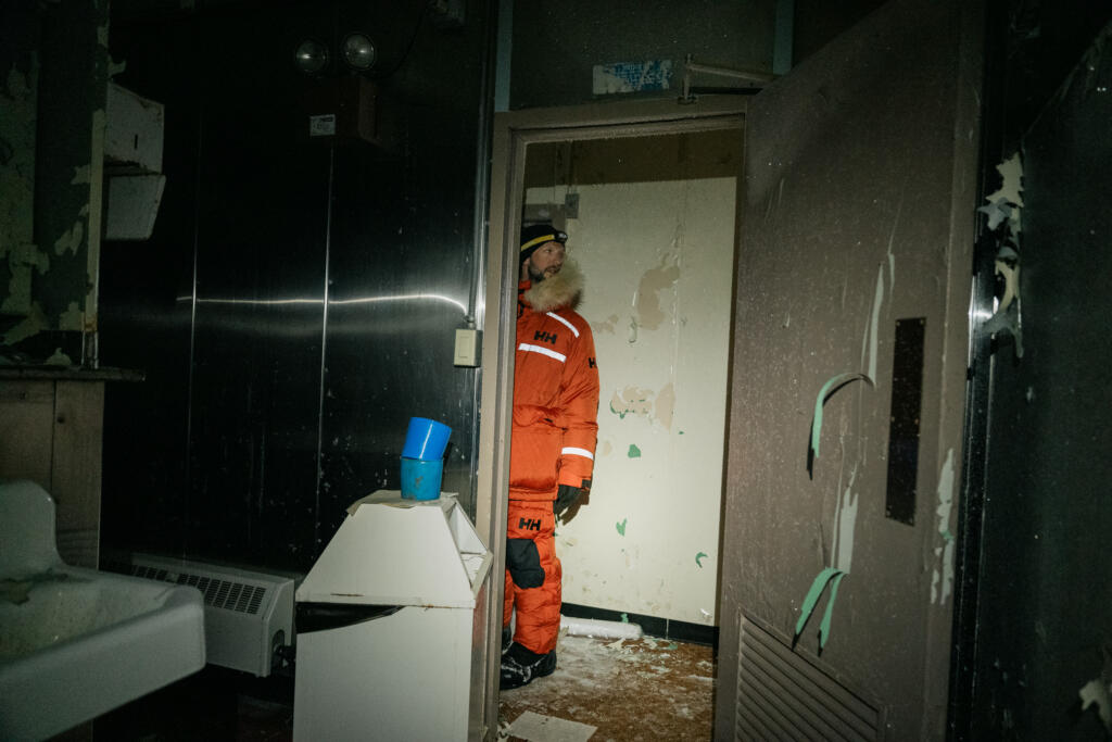 Eine Person in einem orangefarbenen Überlebensanzug steht in einem dunklen Raum mit abblätternder Farbe und verstreuten Gegenständen. Die Tür ist geöffnet, und ein Kühlschrank ist im Hintergrund sichtbar. #Dye-2-greenland, dye-2.