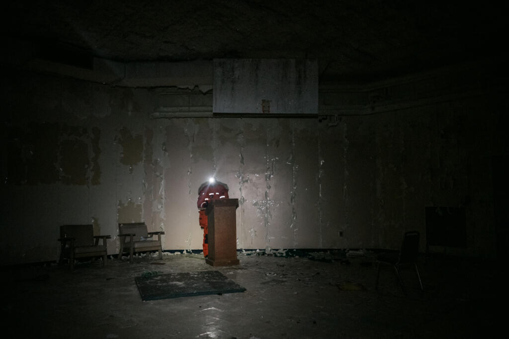 Ein dunkler Raum mit abblätternder Wandfarbe, in dem ein Podium steht, beleuchtet von einer Taschenlampe. Im Hintergrund sind zwei Stühle sichtbar, der Boden ist leer und unordentlich. Die Szene vermittelt eine verlassene Atmosphäre. #Dye-2-greenland