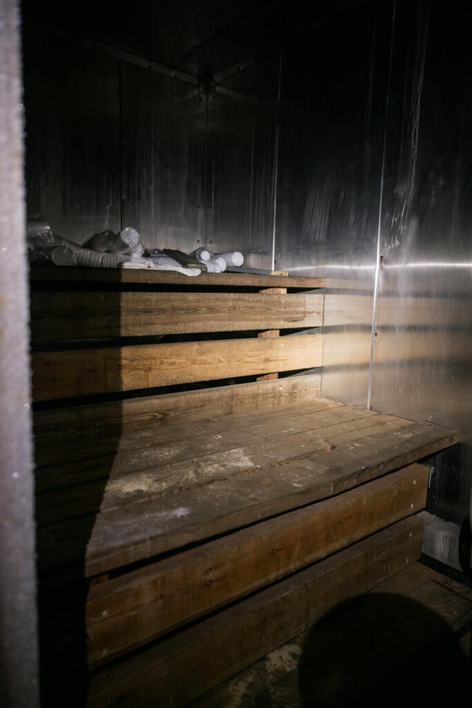 Ein dunkler Raum mit metallischen Wänden und hölzernen Ablagen. Auf den Ablagen liegen einige weiße Objekte. Der Boden ist aus Holz. Die Beleuchtung ist schwach und lässt Schatten entstehen.