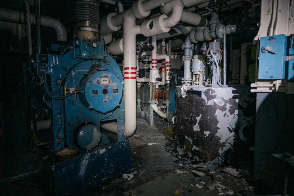 Ein verlassener Maschinenraum mit großen blauen Maschinen und Rohren, die durch die Wände verlaufen. Der Boden ist voller Ablagerungen und die Wände zeigen Schäden. Schwaches Licht beleuchtet den Raum, um einen industriellen Eindruck zu vermitteln. #Dye-2-greenland