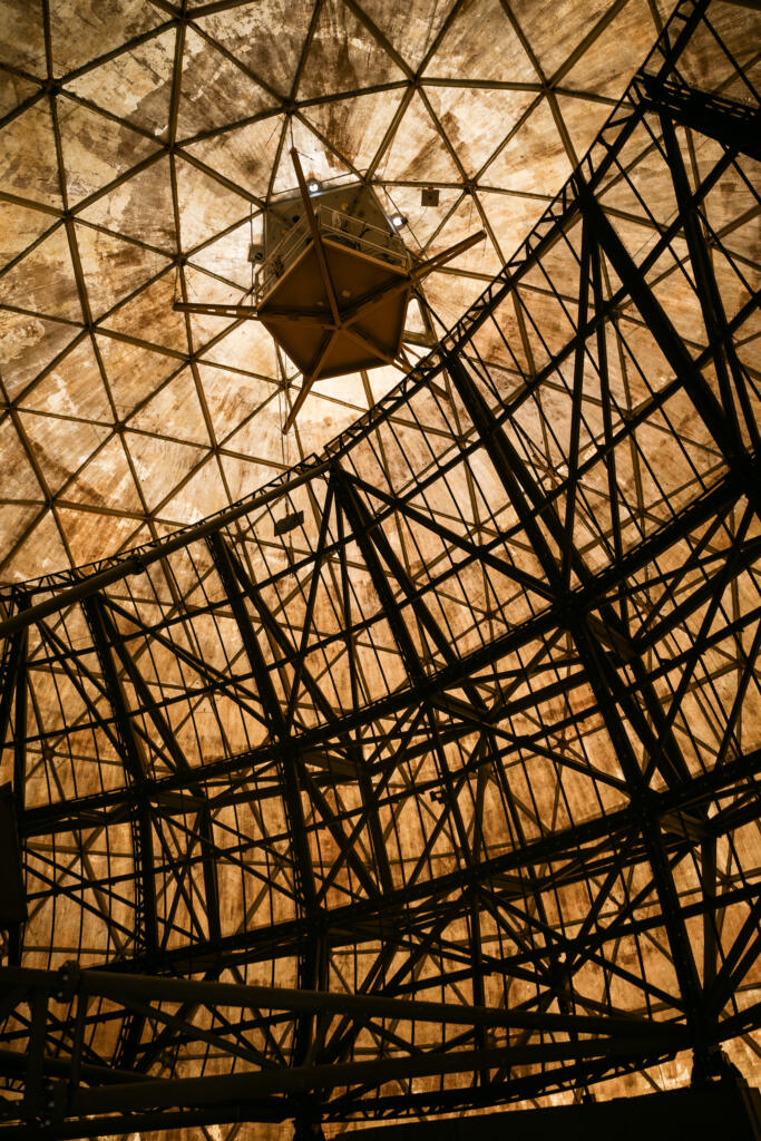 Ein Blick von innen auf die Struktur eines großen, geodätischen Dachs mit einer Metallkonstruktion. Im Mittelpunkt hängt ein geformtes Objekt, während die Oberflächen von Licht beleuchtet werden. Das Bild könnte mit dem Thema #Dye-2-greenland in Verbindung stehen.