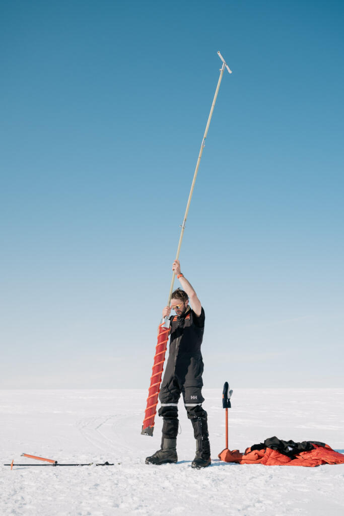 Eine Person in schwarzer Kleidung steht auf einer snow-covered Fläche und hält einen langen Stab mit einer Flagge hoch. Um sie herum ist weißer Schnee sichtbar. Im Hintergrund blauer Himmel ohne Wolken.