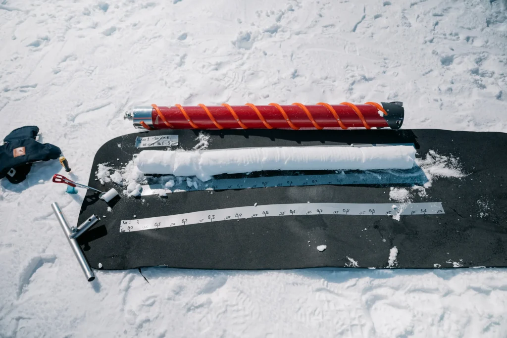 Werkzeuge aus dem Projekt 'The Greenland Project' auf einer Schneefläche, einschließlich eines Schneekerns, einer Bohrmaschine in rot und einem Maßband, die zur Untersuchung des Schnees verwendet werden.