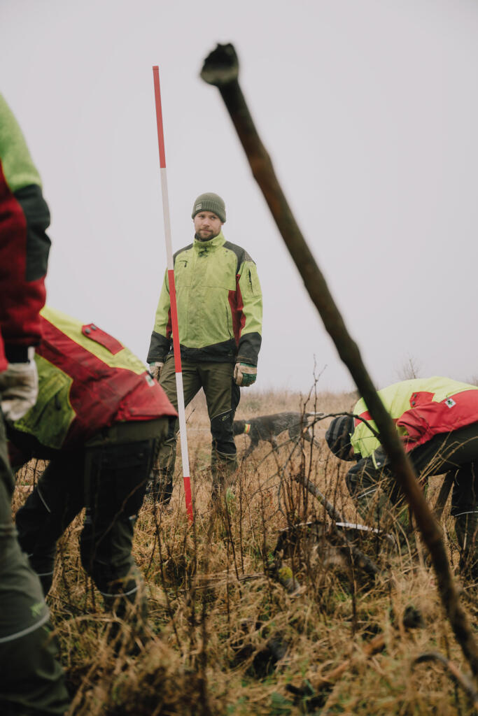 Mehrere Personen, gekleidet in leuchtend gelbe und rote Kleidung, arbeiten in einem grasbewachsenen Gebiet. Eine Person steht in der Mitte mit einem Messstab in der Hand, während andere in der Nähe graben.
