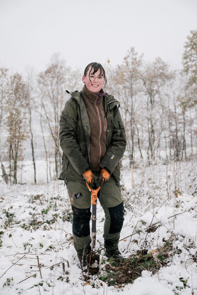 Eine Frau steht im Schnee auf einem bewaldeten Gelände und hält eine Schaufel in der Hand. Sie trägt eine grüne Jacke, einen braunen Pullover und orangefarbene Handschuhe.