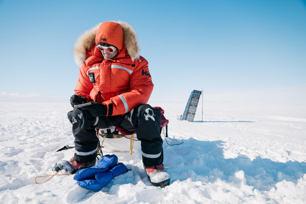 Eine Person in einem orangefarbenen Schneeanzug sitzt auf einem Stuhl in einer schneebedeckten Landschaft und nutzt ein tragbares Gerät. Im Hintergrund ist ein Solarpanel sichtbar.