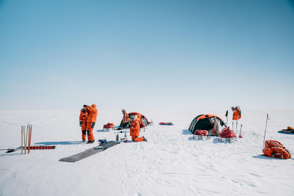 Wissenschaftler in orangen Overalls auf einer schneebedeckten Fläche, umgeben von Zelten und Ausrüstung. Der Himmel ist klar und blau.