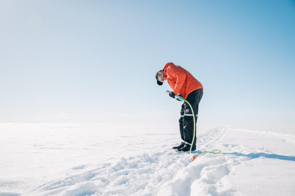 Ein Mann in orangefarbener Jacke beugt sich über den Schnee und hält ein Werkzeug in der Hand, während er sich auf einer schneebedeckten Fläche befindet. Im Hintergrund ist ein klarer blauer Himmel zu sehen. Die Szene ist Teil des Projekts 'The Greenland Project'.