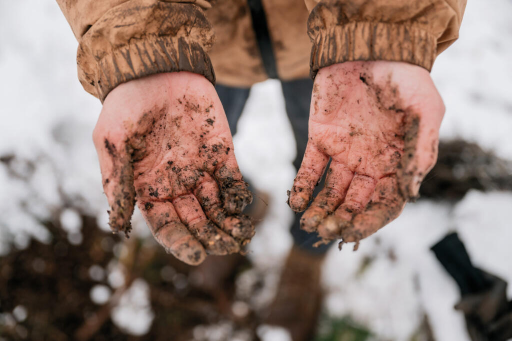 Hände eines Menschen mit Erde und Schmutz, die nach draußen in den Schnee gehalten werden. Im Hintergrund ist eine gefrorene Landschaft sichtbar.
