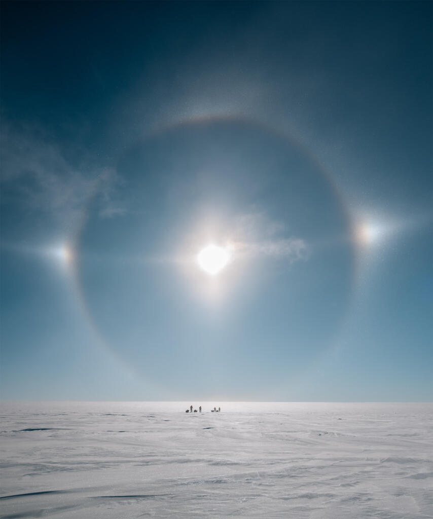 Eine weite, schneebedeckte Landschaft mit einem Sonnenhalo um die Sonne. Im Hintergrund sind kleine Silhouetten von Menschen zu sehen, die sich auf dem gefrorenen Boden bewegen. Der Himmel ist klar und blau.