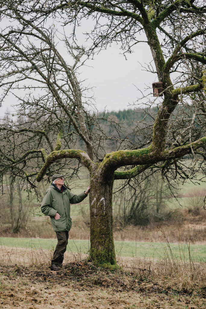 Ein Mann steht neben einem alten, nackten Baum in einer ländlichen Umgebung. Der Baum hat Moos auf den Ästen und eine Vogelbox, die am Stamm angebracht ist. Im Hintergrund sind Wiesen und Wälder sichtbar.