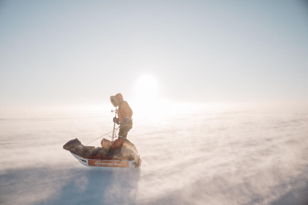 Eine Person zieht einen Schlitten durch eine verschneite Landschaft, während starkes Licht von der Sonne im Hintergrund strahlt. Der Himmel ist klar und die Umgebung wirkt windig mit wirbelndem Schneesturm.
