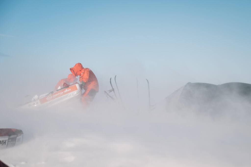 Ein Mensch in orangefarbener Arbeitskleidung steht bei starkem Schneefall und starkem Wind, während er an einem Schneemobil arbeitet. Im Hintergrund sind verschwommene Umrisse von Ausrüstung und Schnee zu sehen.