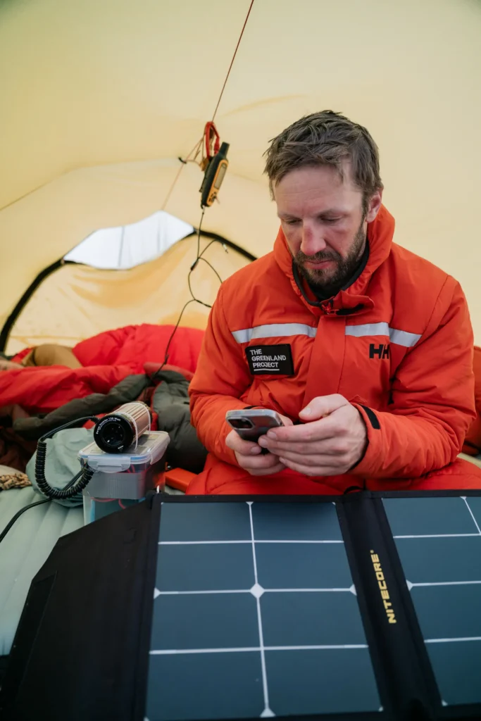 Ein Mann in einem orangefarbenen Anorak sitzt in einem Zelt, während er ein Smartphone benutzt. Vor ihm liegt ein faltbares Solarpanel, und im Hintergrund ist eine Schlafsackausstattung sichtbar. Dieses Bild könnte mit "The Greenland Project" in Verbindung stehen.