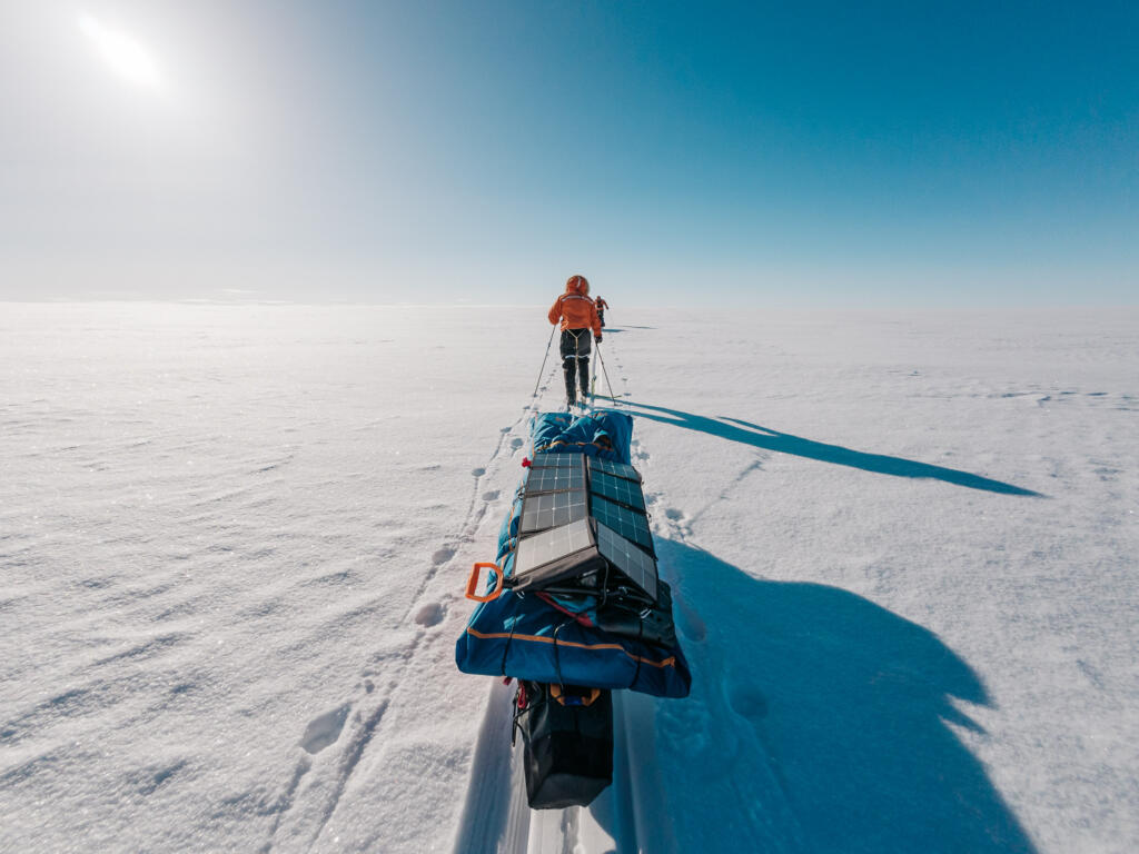 Eine Person in orangefarbener Kleidung zieht einen Schlitten mit modernen Solarpanelen über eine weitläufige, schneebedeckte Fläche unter einem klaren blauen Himmel.