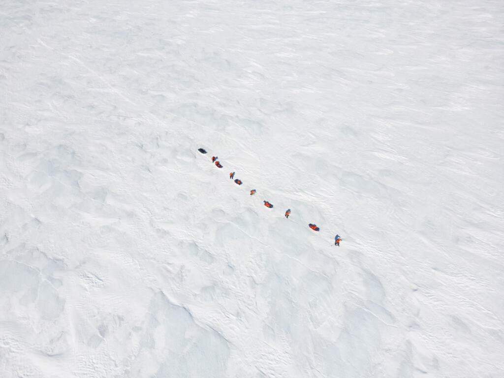 Eine Gruppe von Personen zieht Schlitten über eine weite, schneebedeckte Eisfläche. Die Szene zeigt die Weite der Umgebung und die eisigen Bedingungen.