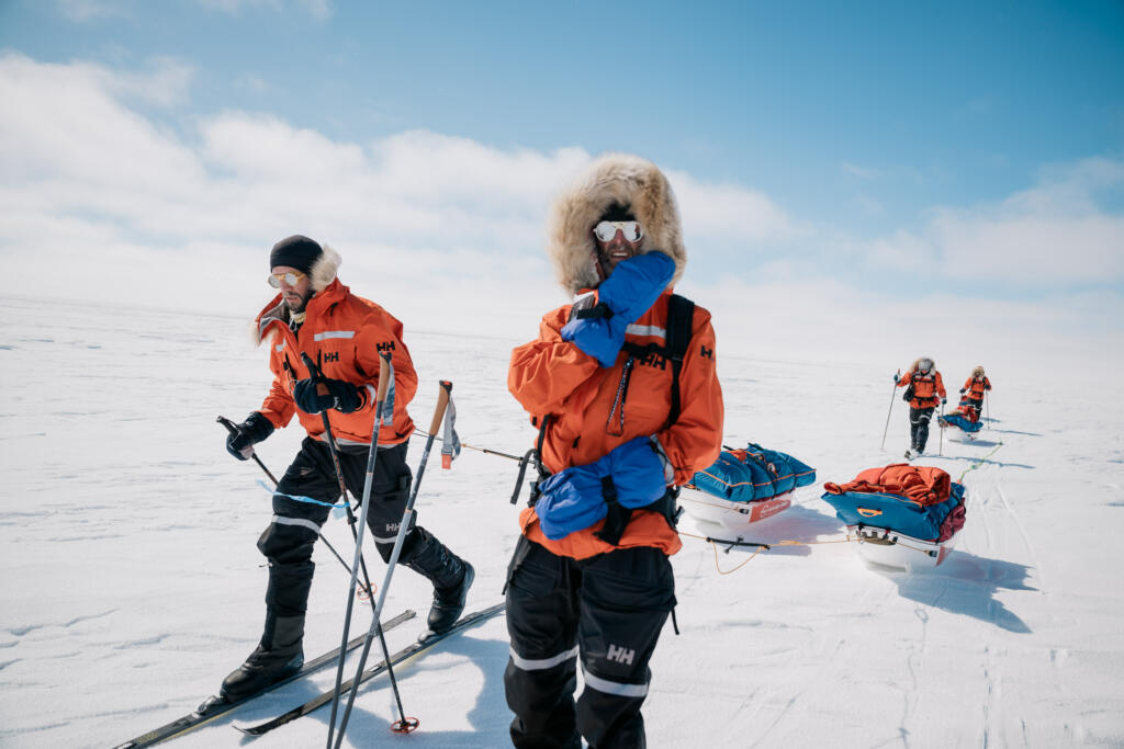 Eine Gruppe von Personen in orange-farbenen Winteranzügen zieht Schlitten über eine schneebedeckte Eisfläche. Einige der Personen tragen Gesichtsschutz und Wärmebekleidung. Der Himmel ist klar mit wenigen Wolken.