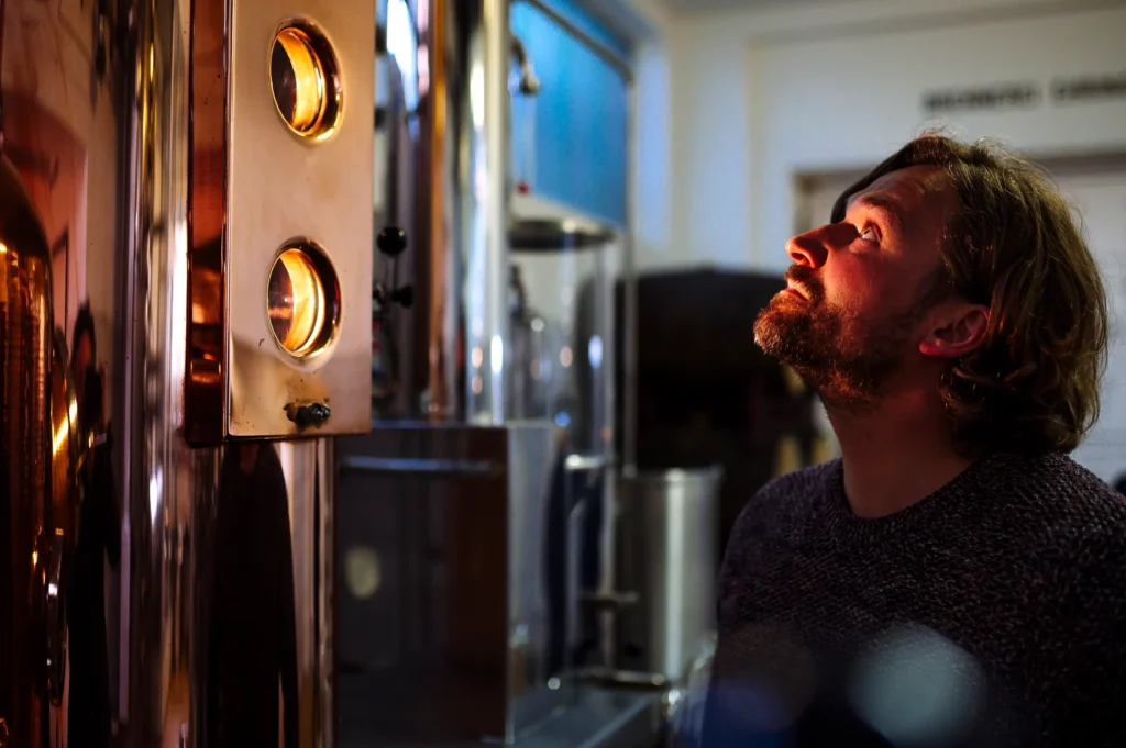 Ein Mann mit lockigem Haar betrachtet aufmerksam eine Brennblase in einer Brennerei. Das Innere der Brennerei zeigt metallische Geräte und eine warme Beleuchtung, die die Szene erhellt. Sichtbar ist auch das Equipment zur Herstellung von Spirituosen der Brennerei Ehringhausen.