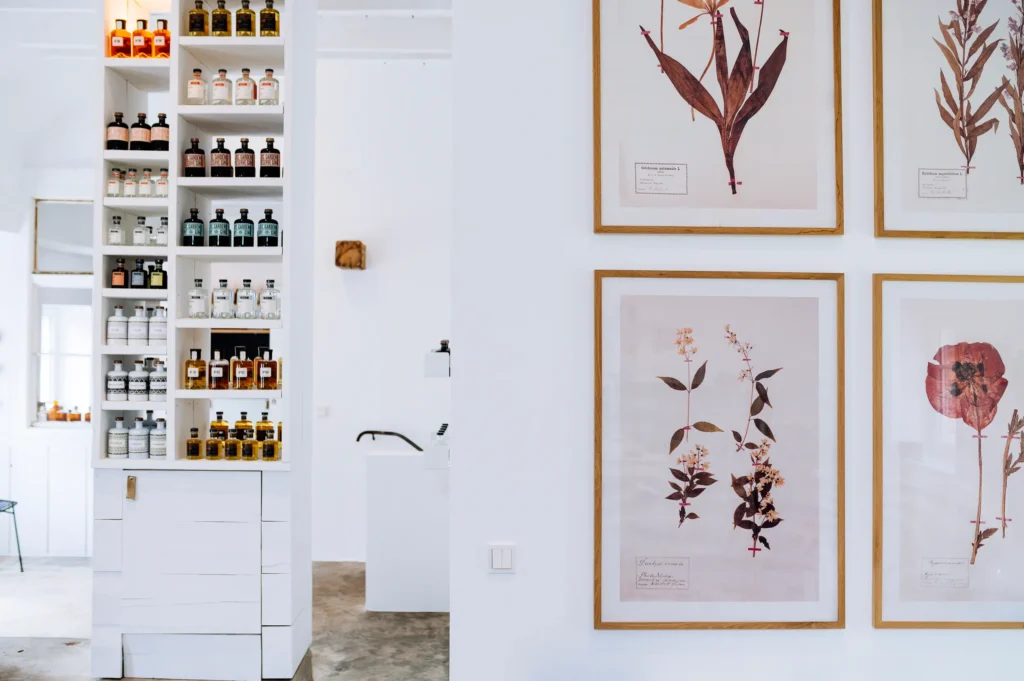 Ein heller Raum mit einer weißen Wand und einer hohen Hyllihe, die Flaschen in verschiedenen Größen zeigt. An der gegenüberliegenden Wand sind gerahmte botanische Drucke ausgestellt. Die Einrichtung ist modern und minimalistisch.