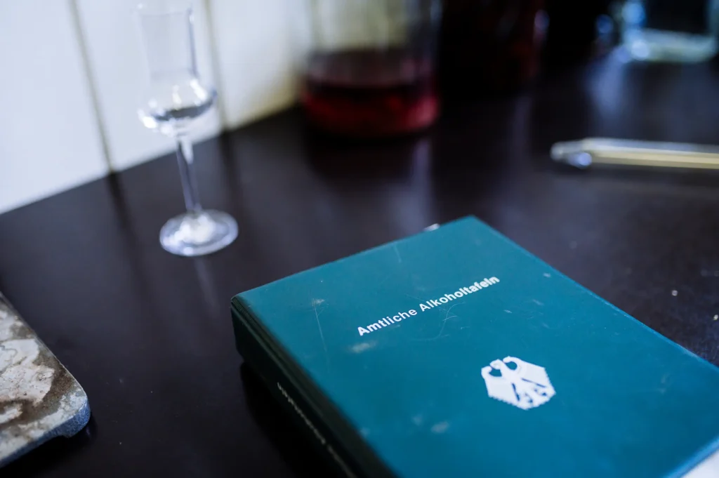 Ein offenes Buch mit dem Titel "Amintices Autohandel" liegt auf einem Tisch neben einem kleinen Glas und einer Flasche. Der Tisch hat eine dunkle Oberfläche, im Hintergrund sind verschwommene Objekte sichtbar. Die Szene könnte mit der Brennerei Ehringhausen in Verbindung stehen.