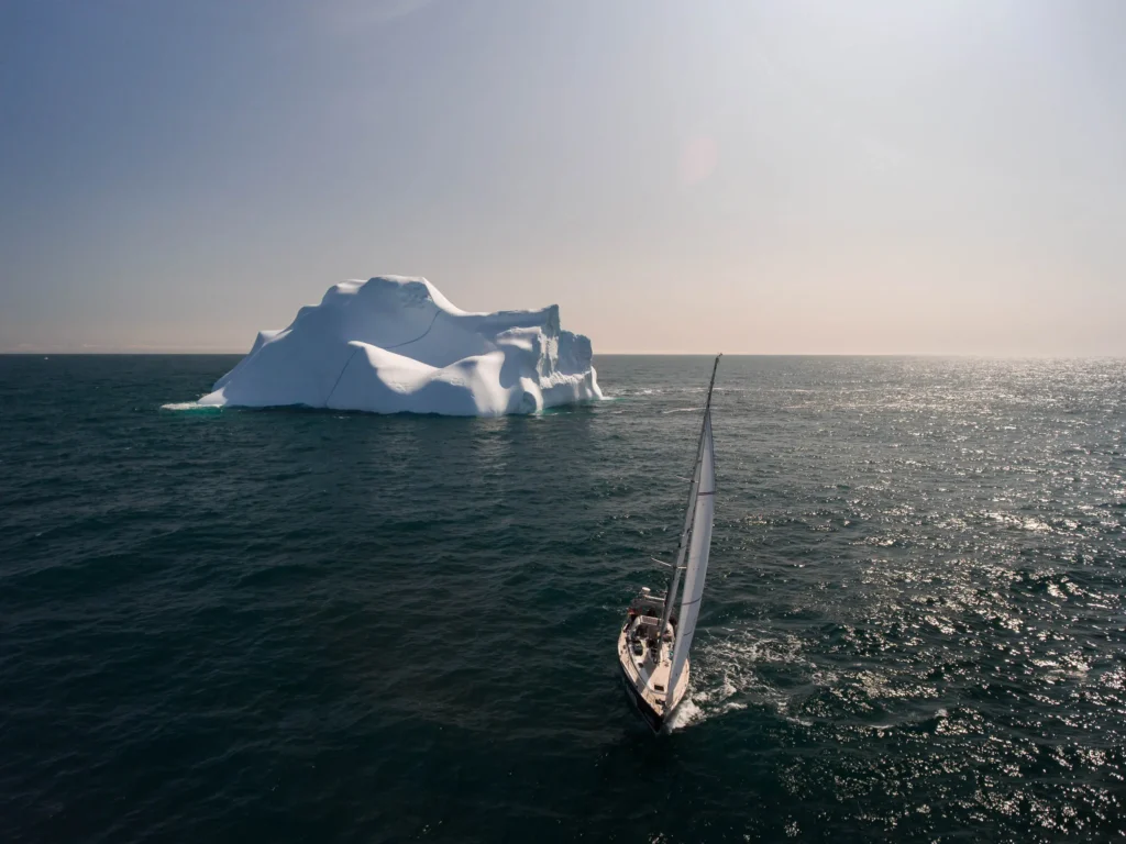 Ein Segelboot fährt in der Nähe eines großen Eisbergs auf ruhiger See mit Sonnenschein und klarem Himmel. Die Wellen reflektieren das Licht und schaffen eine ruhige maritime Szene. Keyword: geo saison.