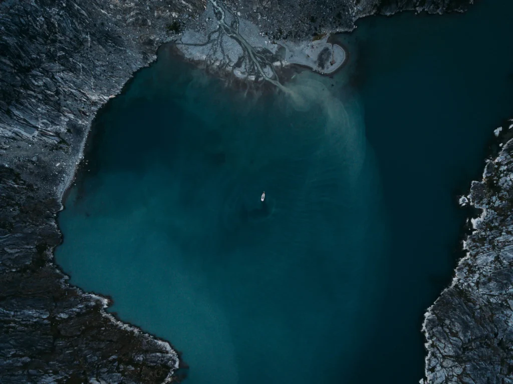 Luftaufnahme eines Gewässers mit tiefblauem Wasser, umgeben von felsigen Küsten und kleinen Kiesstränden. Ein einzelnes Boot befindet sich auf dem Wasser, umgeben von der ruhigen Natur der Umgebung. Die Aufnahme zeigt geografische Merkmale des Gebiets, passend für die Thematisierung von "geo saison".