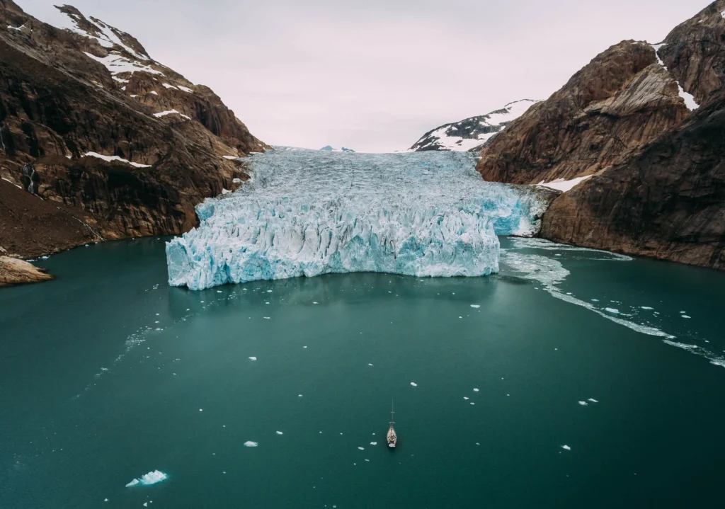Ein Schiff fährt in einer Fjordlandschaft mit einem großen Gletscher und schneebedeckten Bergen. Eisschollen treiben im Wasser, während der Himmel bewölkt ist. Die Szene vermittelt eine majestätische, natürliche Umgebung.