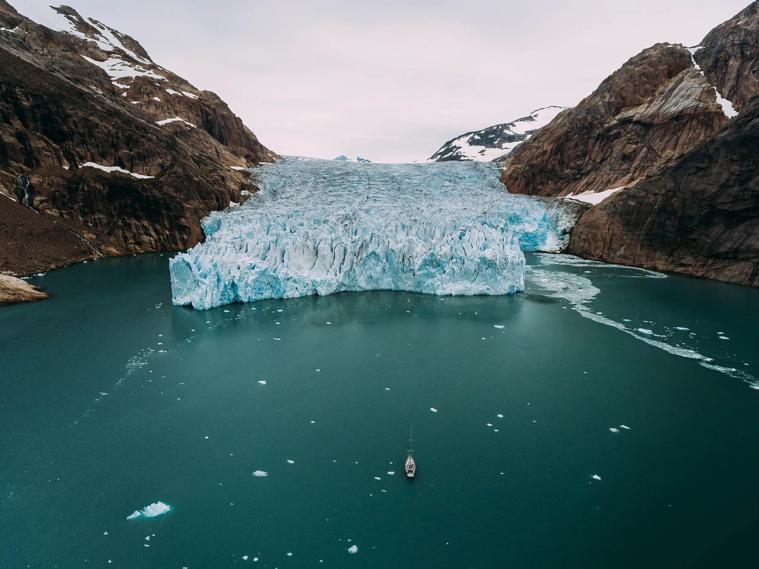 Eine Nahaufnahme eines großen Gletschers, der in ein blaues Wasserbecken kalbt, umgeben von steilen Felsen und schneebedeckten Bergen. Ein kleines Boot befindet sich in der Nähe des Gletschers. Segelfotografie.