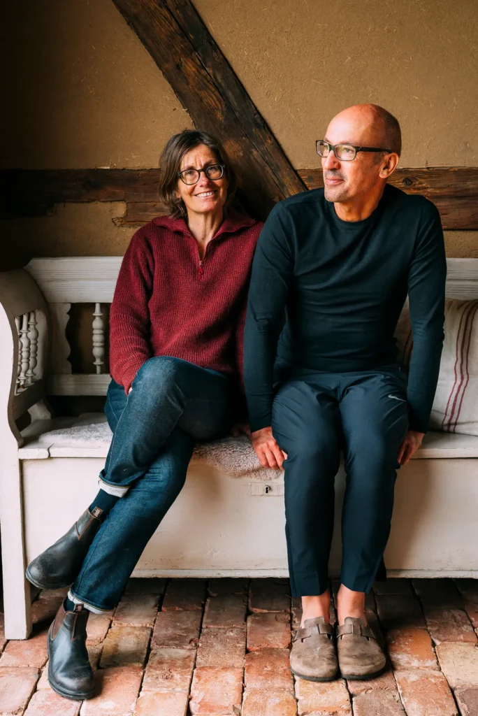 Ein Paar sitzt auf einer Bank in einem Raum mit Holzbalken. Die Frau trägt einen roten Pullover und hat eine Brille, der Mann trägt ein schwarzes Oberteil. Der Boden ist mit alten Ziegelsteinen bedeckt. Sie scheinen entspannt zu sein.