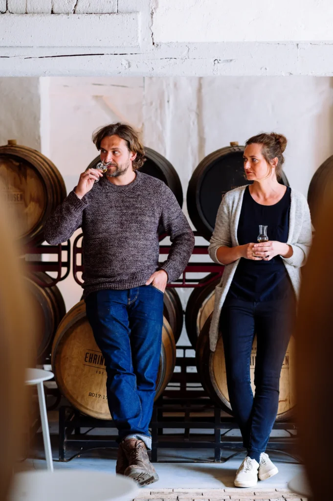 Ein Mann und eine Frau stehen in einer Weinlagerung, umgeben von Holzfässern. Der Mann hält ein Glas und schaut nachdenklich, während die Frau in einem entspannten Outfit ein weiteres Glas in der Hand hält.