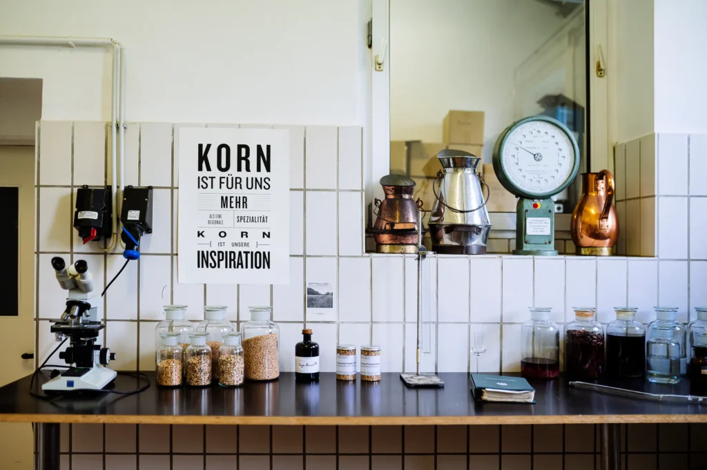 Eine Küchenzeile mit verschiedenen Utensilien, darunter ein Mikroskop, mehrere Glasbehälter mit Körnern, eine Kaffeekanne und ein Wandzeitmesser. An der Wand hängt ein Poster mit der Aufschrift: "KORN IST FÜR UNS MEHR INSPIRATION". Diese Szene befindet sich in einer Brennerei Ehringhausen.
