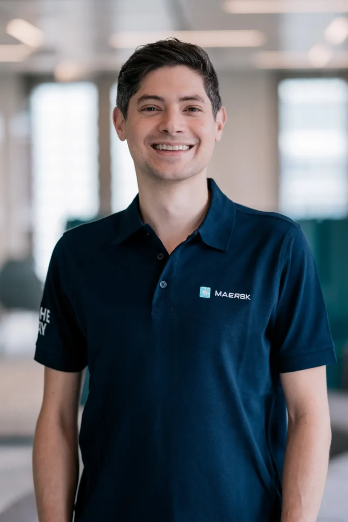 Ein Mann in einem blauen Poloshirt mit dem Logo von Maersk steht in einem modernen Bürogebäude und lächelt in die Kamera.