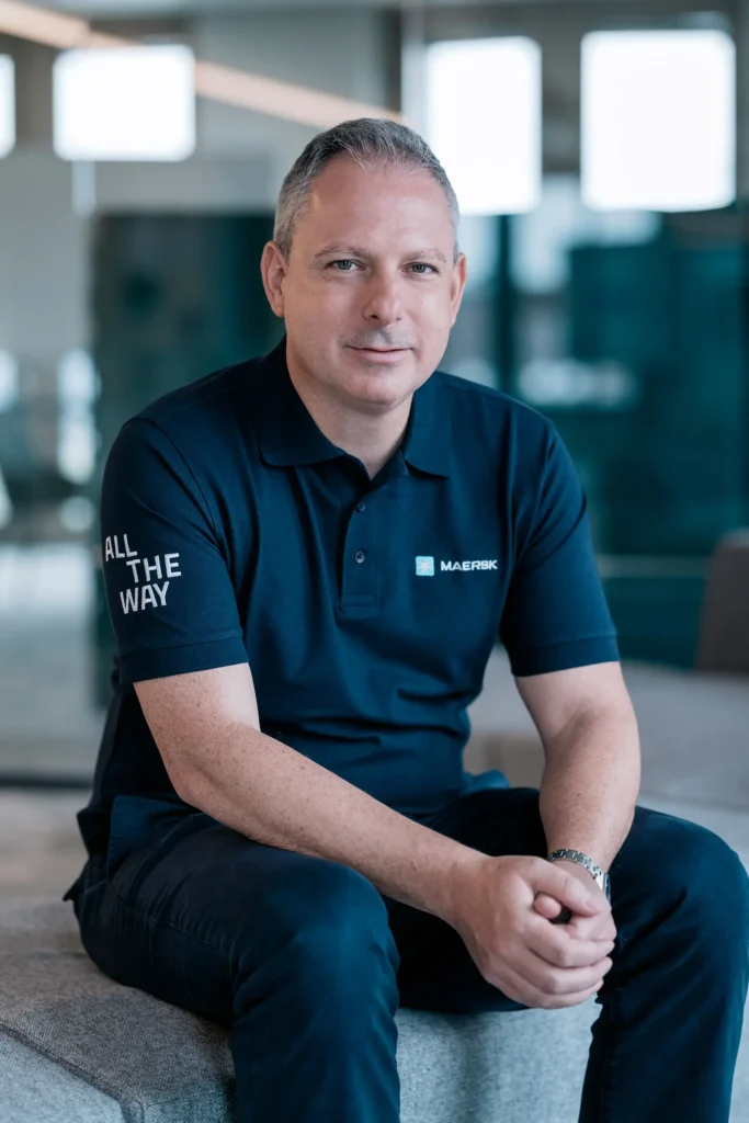 Ein Mann sitzt auf einer Couch in einem modernen Büro, trägt ein schwarzes Poloshirt mit der Aufschrift "ALL THE WAY" und einem Logo. Er hat einen neutralen Ausdruck und sieht direkt in die Kamera.