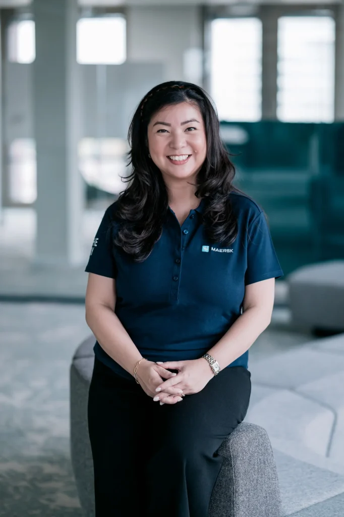Eine Frau mit langen, dunkelbraunen Haaren sitzt lächelnd auf einer grauen Couch in einem modernen Büro. Sie trägt ein dunkelblaues Polo-Shirt mit einem Logo und hat eine entspannte Haltung.