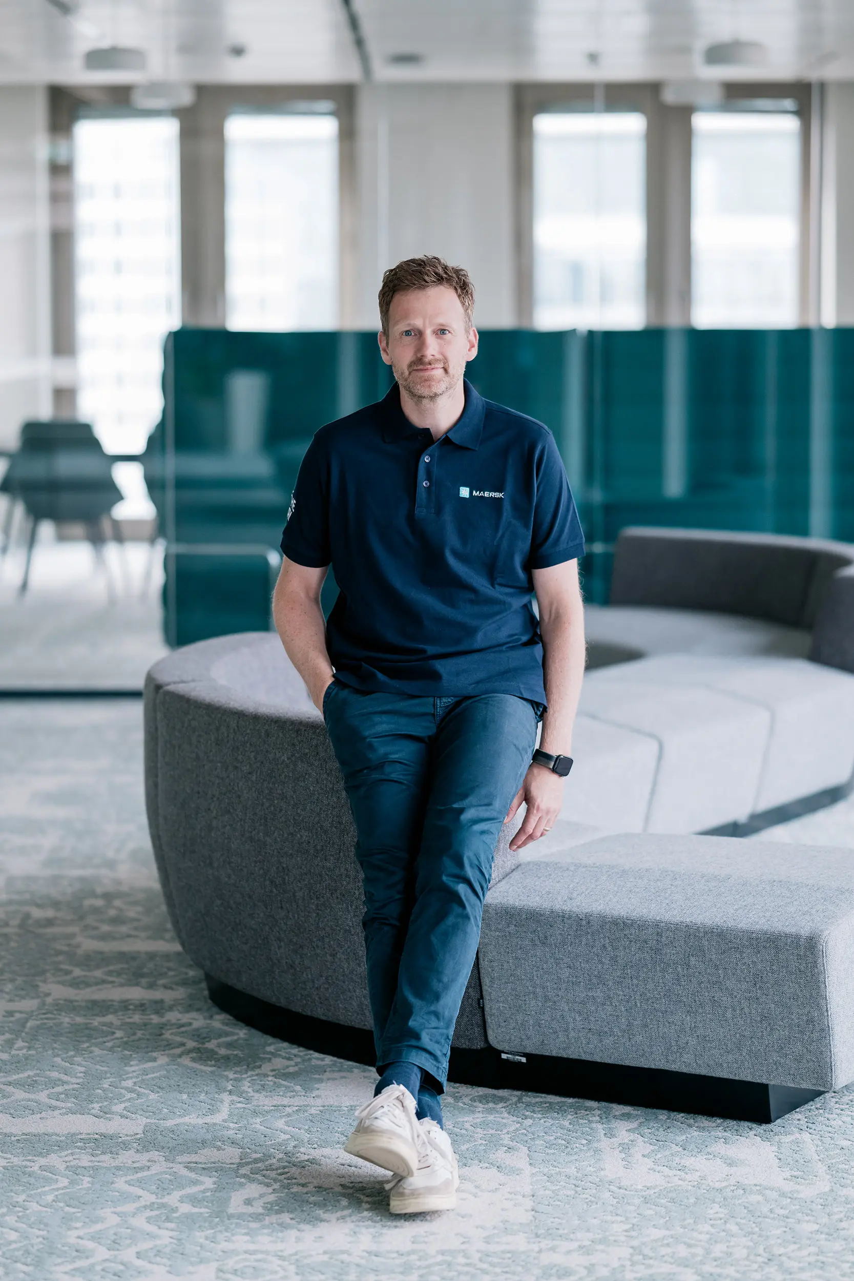 Ein Mann in einem blauen Polohemd und Jeans steht lässig an einer grauen Couch in einem modernen Büro mit großen Fenstern im Hintergrund.