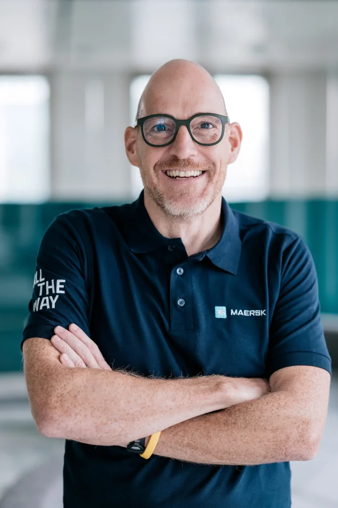 Ein Mann mit Glatze und Brille lächelt in einem modernen Büro. Er trägt ein dunkelblaues Poloshirt mit dem Logo von Maersk. Die Umgebung ist hell und freundlich gestaltet.
