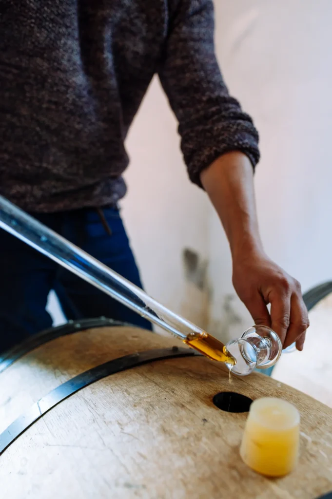 Ein Mann verwendet eine Pipette, um eine goldene Flüssigkeit aus einem Weinfass in ein Glas zu gießen. Im Vordergrund steht ein Wachszylinder. Die Szene zeigt einen Arbeitsbereich in einer Brennerei Ehringhausen.