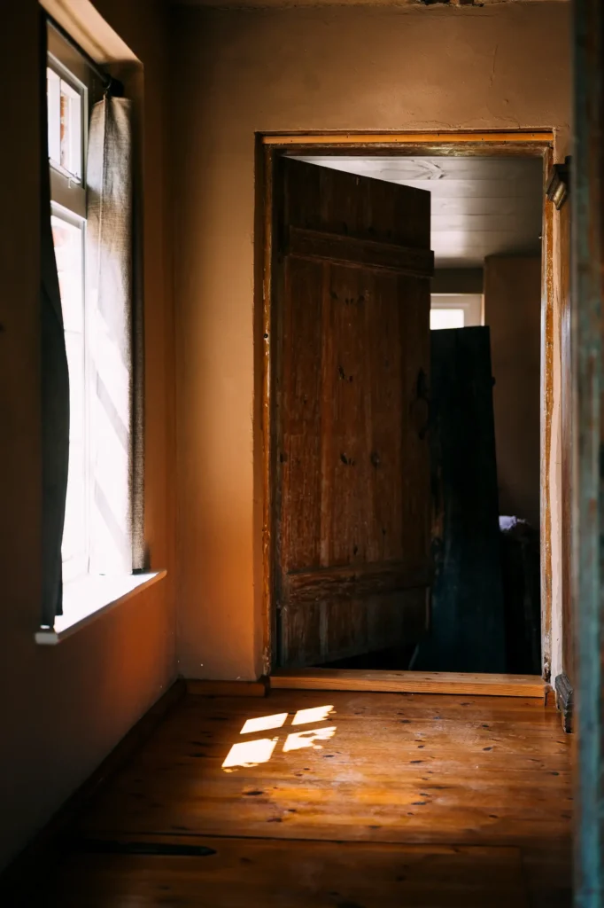 Ein heller Flur mit Lichtstrahlen, die durch ein Fenster auf den Holzboden fallen. Eine geöffnete Holztür führt in einen weiteren Raum. Die Wände sind schlicht und in neutralen Tönen gehalten. Diese Szene könnte Teil einer "meine kleine farm" oder "wildyard" Umgebung sein.