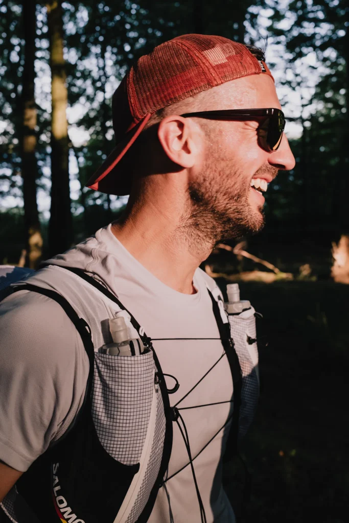 Ein Mann mit einer roten Kappe und Sonnenbrille steht lächelnd im Wald, während er einen Rucksack mit Trinkflaschen trägt. Sonnenlicht fällt durch die Bäume und beleuchtet die Szene.