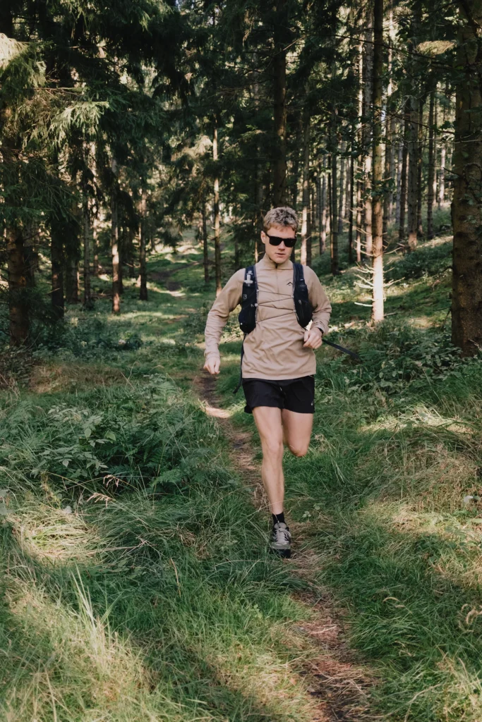Ein Mann in Sportkleidung mit Rucksack joggt auf einem Pfad durch einen Wald, umgeben von hohen Bäumen und üppigem Gras.