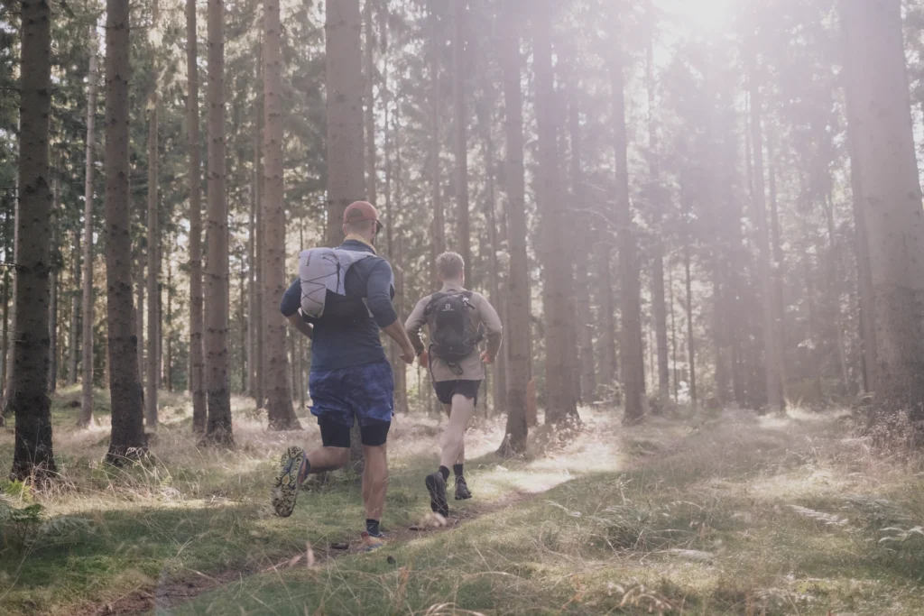 Zwei Männer in Sportkleidung joggen auf einem schmalen Weg durch einen Wald mit hohen Bäumen. Die Sonne scheint durch die Baumkronen.
